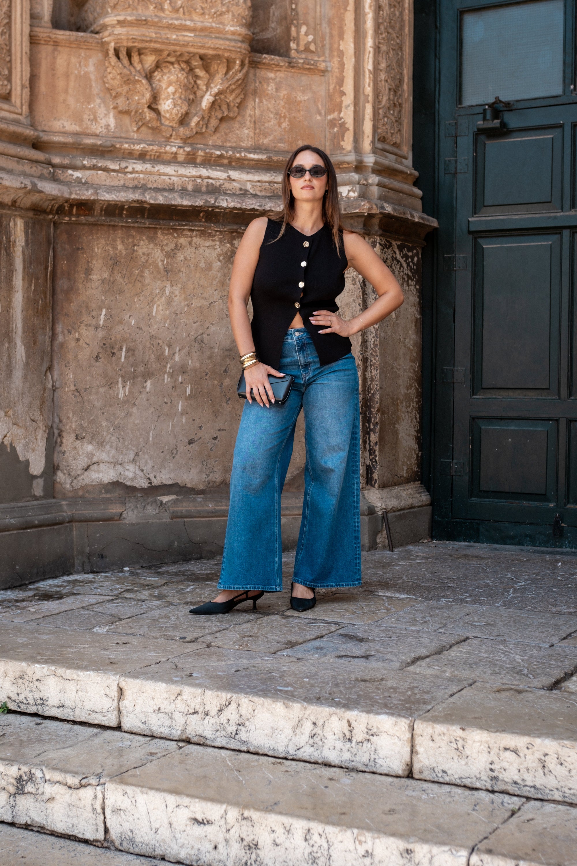 Woman in black top and blue jeans standing in front of a stone building.