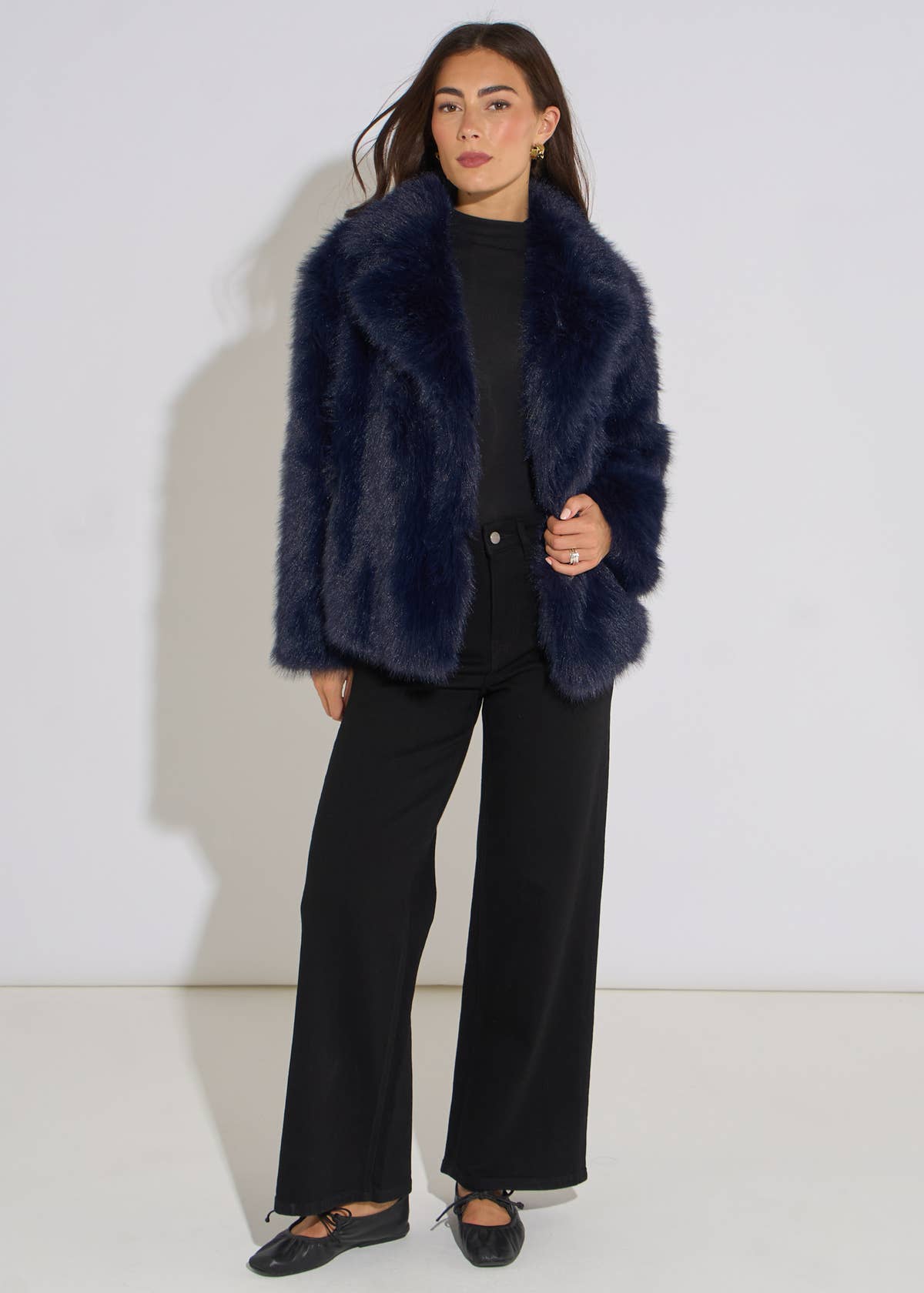 A woman modeling a dark navy blue shaggy faux fur jacket over a black top and black wide-leg trousers, standing against a plain white studio background.